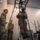 Leapfest 2026 (Pre Jump Training) Military personnel are conducting what appears to be a parachute or airborne training simulation inside an indoor facility. One soldier wearing a VR headset and harness is suspended from an overhead rigging system, while two other soldiers in US Army OCP uniforms observe. The setup suggests a virtual reality-assisted jump training or egress simulation exercise.
