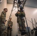Military personnel are conducting what appears to be a parachute or airborne training simulation inside an indoor facility. One soldier wearing a VR headset and harness is suspended from an overhead rigging system, while two other soldiers in US Army OCP uniforms observe. The setup suggests a virtual reality-assisted jump training or egress simulation exercise.