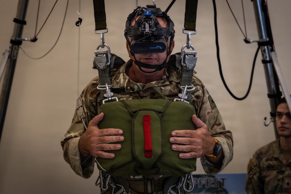 A military personnel member is suspended in a parachute training harness rig while wearing a virtual reality headset, simulating a parachute jump experience. The individual is dressed in multicam/MTP camouflage uniform and is gripping what appears to be a simulated parachute reserve handle. This appears to be a military parachute training simulator combining VR technology with physical harness equipment.