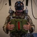 A military personnel member is suspended in a parachute training harness rig while wearing a virtual reality headset, simulating a parachute jump experience. The individual is dressed in multicam/MTP camouflage uniform and is gripping what appears to be a simulated parachute reserve handle. This appears to be a military parachute training simulator combining VR technology with physical harness equipment.