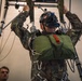 A military personnel wearing camouflage uniform is suspended in a parachute training harness rig while wearing a virtual reality headset with additional mounted equipment. The setup appears to be an indoor VR-enhanced parachute simulation trainer, combining physical harness suspension with immersive virtual reality technology. A second individual in camouflage is partially visible in the background.