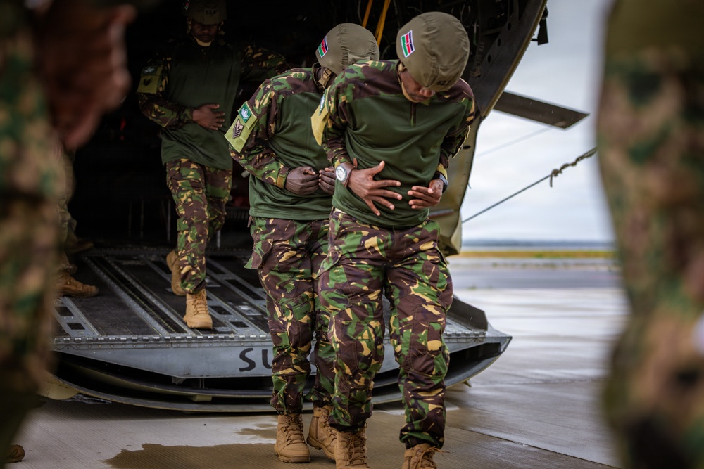 Military personnel wearing camouflage uniforms with what appears to be Kenyan flag patches are boarding or exiting a military helicopter via its rear ramp. Two soldiers in the foreground appear to be assisting or steadying each other near the ramp. The setting appears to be a wet tarmac or airfield under overcast conditions.