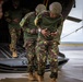 Military personnel wearing camouflage uniforms with what appears to be Kenyan flag patches are boarding or exiting a military helicopter via its rear ramp. Two soldiers in the foreground appear to be assisting or steadying each other near the ramp. The setting appears to be a wet tarmac or airfield under overcast conditions.