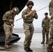 Military personnel in multicam camouflage uniforms are gathered near the rear ramp of a large military helicopter or transport aircraft on a tarmac. Several soldiers appear to be conducting pre-mission or post-mission equipment checks, with two wearing helmets with netting. The setting appears to be an airfield under overcast skies.