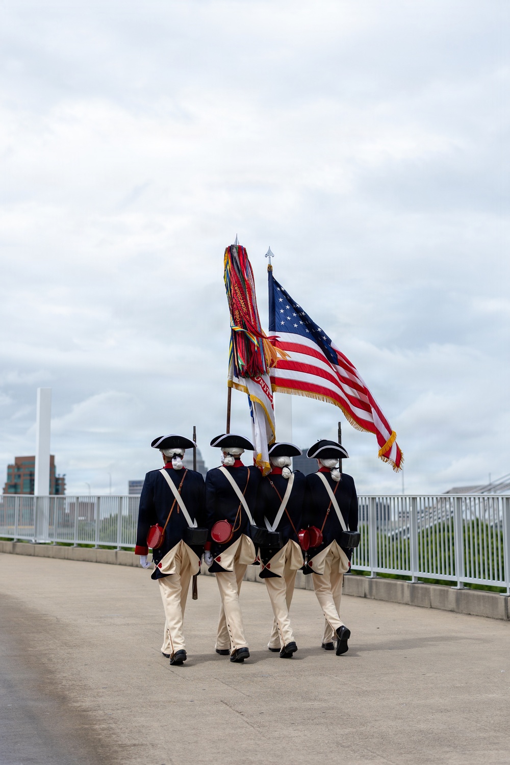 Four individuals dressed in American Revolutionary War-era military uniforms are marching in formation on a paved walkway or bridge, carrying the United States flag and at least one additional ceremonial flag. They are wearing tricorn hats, dark blue coats, cream-colored breeches, white cross-body sashes, and face masks, suggesting this is a color guard ceremony during a period requiring COVID-19 precautions. An urban skyline is partially visible in the background.