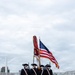 Four individuals dressed in American Revolutionary War-era military uniforms are marching in formation on a paved walkway or bridge, carrying the United States flag and at least one additional ceremonial flag. They are wearing tricorn hats, dark blue coats, cream-colored breeches, white cross-body sashes, and face masks, suggesting this is a color guard ceremony during a period requiring COVID-19 precautions. An urban skyline is partially visible in the background.