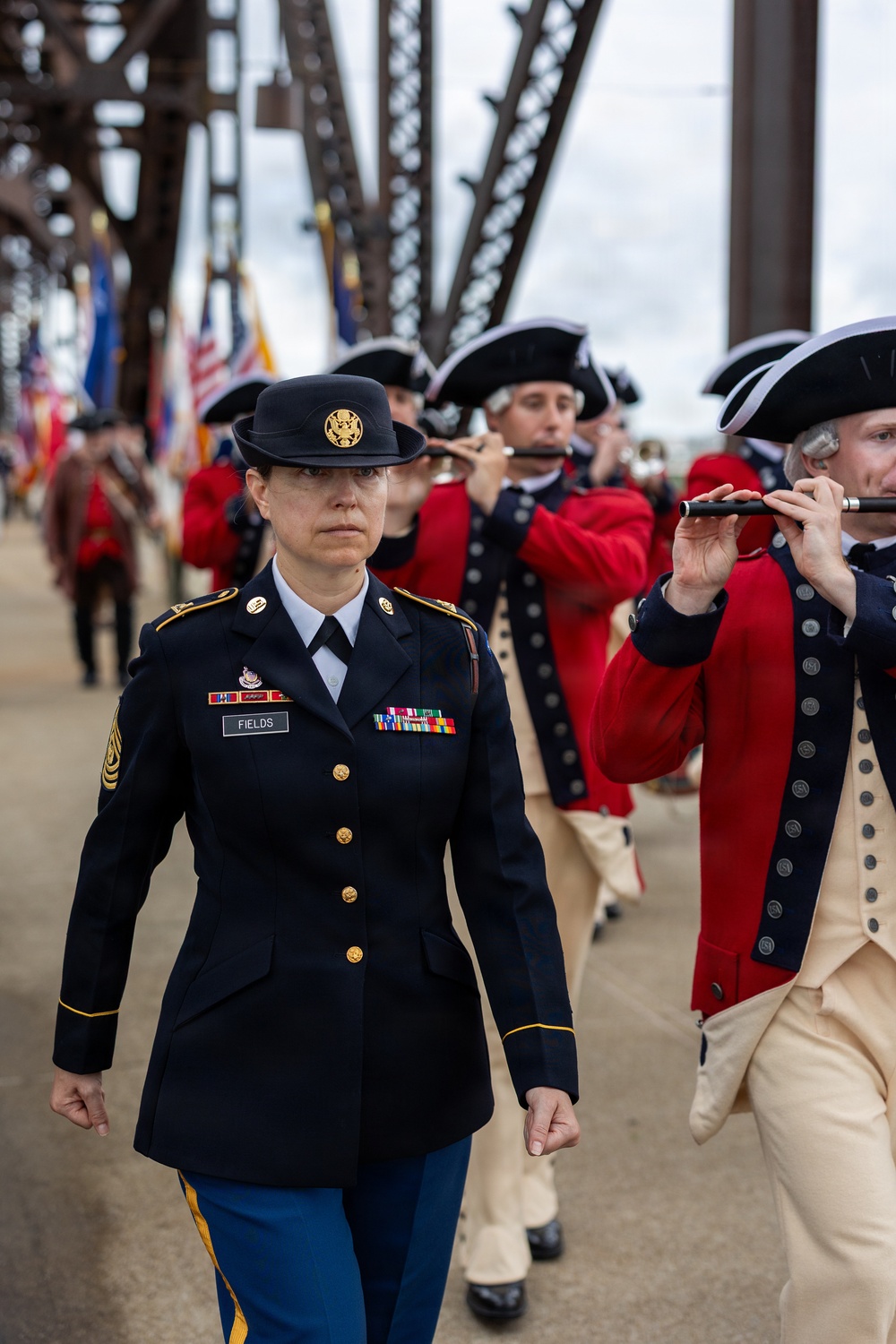 A U.S. Army soldier named Fields, bearing the rank of Sergeant Major, leads a ceremonial military parade across what appears to be a steel truss bridge. Behind her, musicians dressed in 18th-century Continental Army-style uniforms (red coats and tricorn hats) play fifes and flutes. Color guard members carrying American and military unit flags are visible in the background.