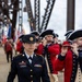 A U.S. Army soldier named Fields, bearing the rank of Sergeant Major, leads a ceremonial military parade across what appears to be a steel truss bridge. Behind her, musicians dressed in 18th-century Continental Army-style uniforms (red coats and tricorn hats) play fifes and flutes. Color guard members carrying American and military unit flags are visible in the background.