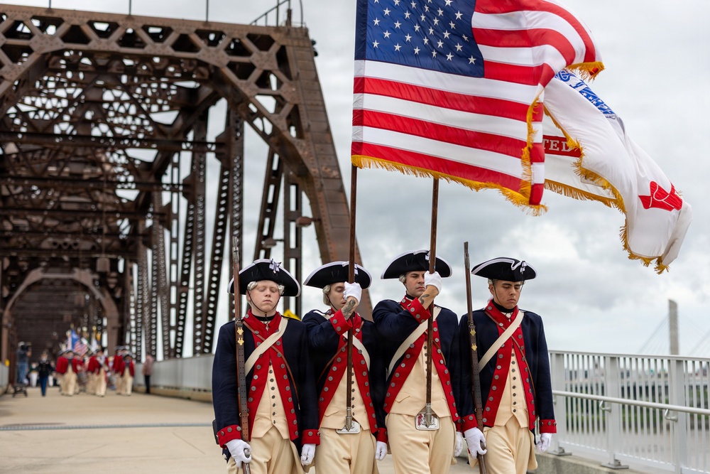 A color guard unit dressed in 18th-century Continental Army-style uniforms marches across a large iron truss bridge carrying the American flag and at least one additional military or organizational flag. The participants wear tricorn hats, navy and red coats with cream-colored breeches, white gloves, and carry period-style muskets. This appears to be a ceremonial parade or patriotic event, likely associated with a military commemoration or national holiday.
