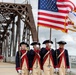A color guard unit dressed in 18th-century Continental Army-style uniforms marches across a large iron truss bridge carrying the American flag and at least one additional military or organizational flag. The participants wear tricorn hats, navy and red coats with cream-colored breeches, white gloves, and carry period-style muskets. This appears to be a ceremonial parade or patriotic event, likely associated with a military commemoration or national holiday.