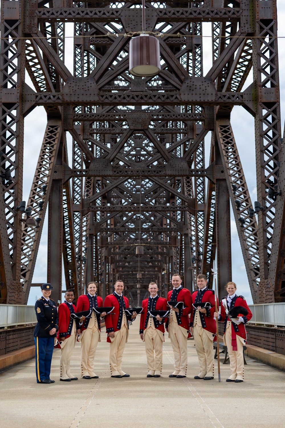 A group of military personnel pose on a large steel truss bridge, with most members wearing 18th-century style ceremonial uniforms consisting of red coats, cream breeches, and tricorn hats. One individual on the far left wears a modern U.S. Army service uniform. The bridge's riveted steel lattice framework creates a dramatic symmetrical backdrop receding into the distance.