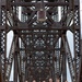 A group of military personnel pose on a large steel truss bridge, with most members wearing 18th-century style ceremonial uniforms consisting of red coats, cream breeches, and tricorn hats. One individual on the far left wears a modern U.S. Army service uniform. The bridge's riveted steel lattice framework creates a dramatic symmetrical backdrop receding into the distance.