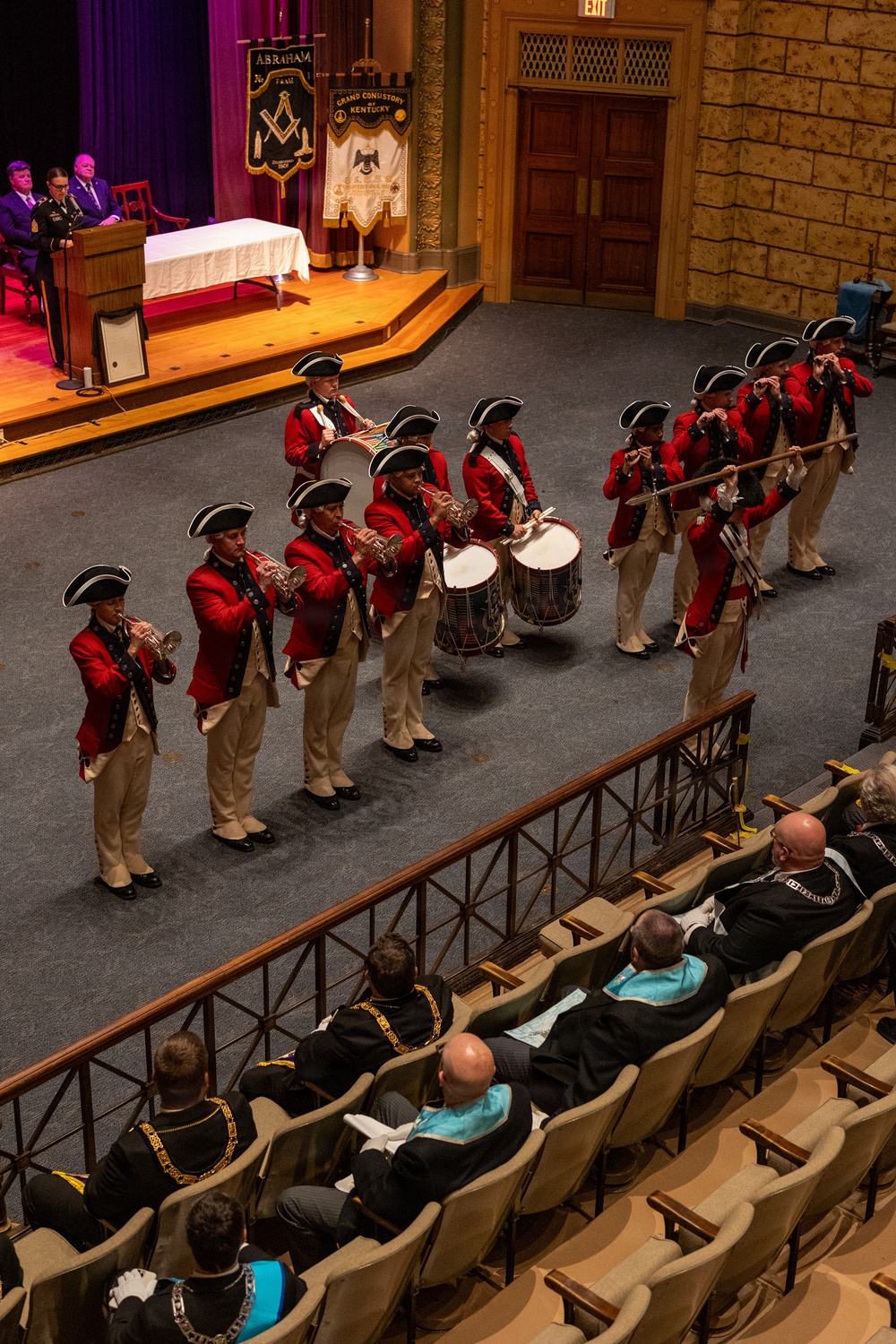 A colonial-era style fife and drum corps in red coats and tricorn hats performs in an auditorium before a seated audience. The performers are arranged in formation playing bugles/trumpets, drums, and fifes. The event appears to be a Masonic ceremony, evidenced by banners reading 'Grand Consistory of Kentucky' and audience members wearing Masonic regalia.