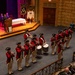 A colonial-era style fife and drum corps in red coats and tricorn hats performs in an auditorium before a seated audience. The performers are arranged in formation playing bugles/trumpets, drums, and fifes. The event appears to be a Masonic ceremony, evidenced by banners reading 'Grand Consistory of Kentucky' and audience members wearing Masonic regalia.