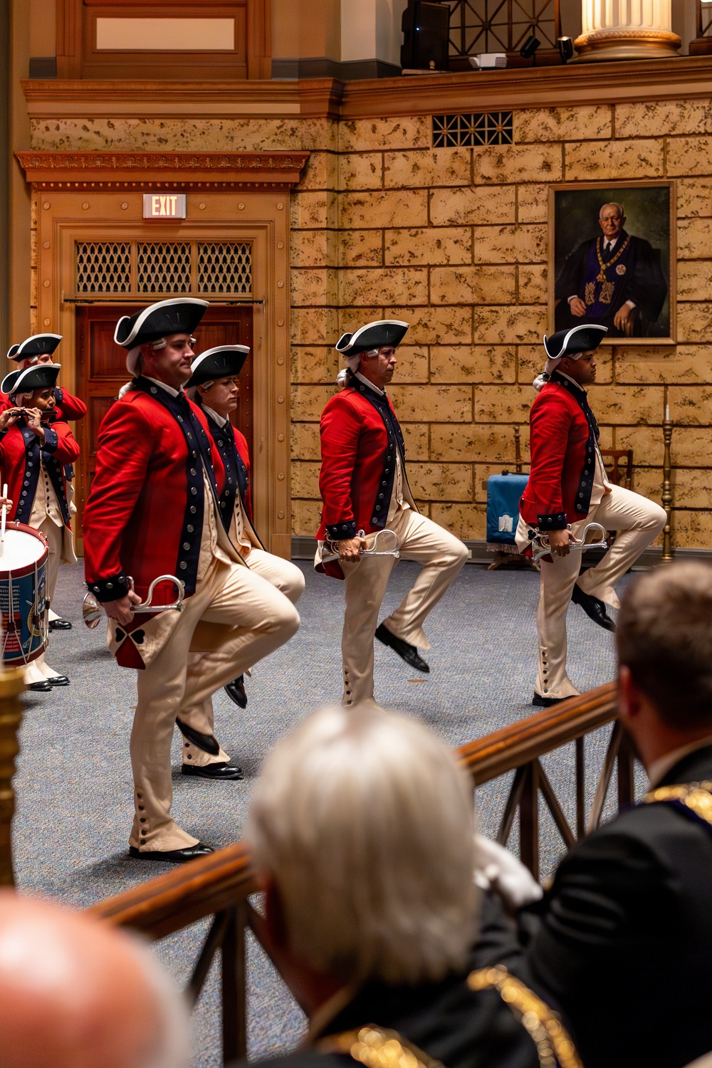 A group of performers dressed in 18th-century American Revolutionary War-era military uniforms — red coats, tricorn hats, white wigs, and cream breeches — are marching in formation inside an ornate indoor hall. They carry swords at their sides and perform a high-stepping march while a drummer and fifer accompany them. The setting appears to be a formal ceremonial event, likely at a Masonic or fraternal organization hall, as evidenced by the portrait on the wall depicting a figure in Masonic regalia.