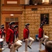 A group of performers dressed in 18th-century American Revolutionary War-era military uniforms — red coats, tricorn hats, white wigs, and cream breeches — are marching in formation inside an ornate indoor hall. They carry swords at their sides and perform a high-stepping march while a drummer and fifer accompany them. The setting appears to be a formal ceremonial event, likely at a Masonic or fraternal organization hall, as evidenced by the portrait on the wall depicting a figure in Masonic regalia.
