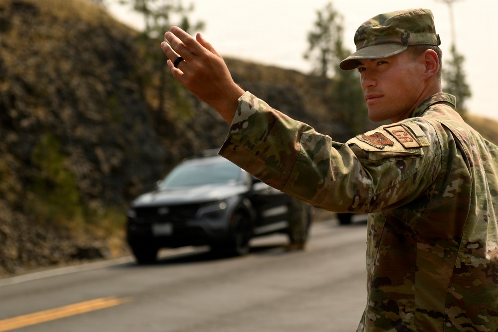 A military service member in MultiCam/OCP camouflage uniform is directing traffic on a two-lane road, raising his hand in a stop or halt gesture toward an approaching dark-colored vehicle. The setting appears to be a rural or semi-arid area with rocky terrain and sparse trees in the background. The scene suggests a traffic control point or checkpoint operation.