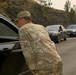Military personnel in OCP (Operational Camouflage Pattern) uniforms are conducting a vehicle checkpoint or traffic control operation on a road flanked by rocky terrain and pine trees. At least two soldiers are visible, one leaning into the window of a black Jeep Grand Cherokee in the foreground, and another further down the line interacting with additional vehicles. This appears to be a security checkpoint, evacuation control point, or access restriction operation.