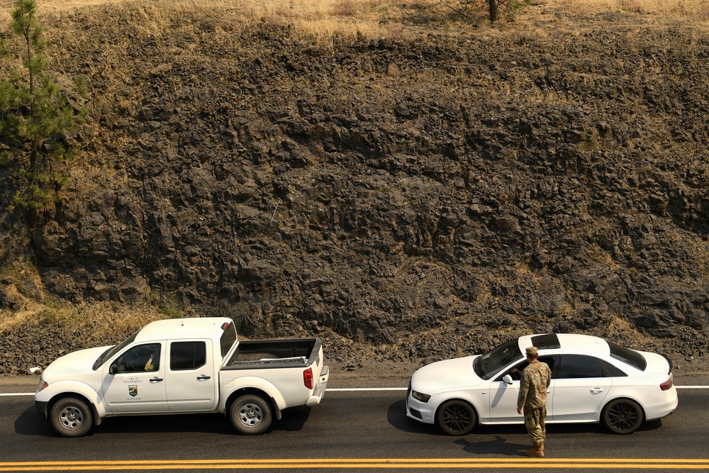 An aerial or elevated view of two white vehicles stopped on a paved road alongside a rocky cliff face. A person in camouflage military or law enforcement attire is standing between the vehicles, appearing to interact with the occupant of a white sedan. The scene suggests a checkpoint or traffic stop situation.