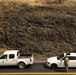 An aerial or elevated view of two white vehicles stopped on a paved road alongside a rocky cliff face. A person in camouflage military or law enforcement attire is standing between the vehicles, appearing to interact with the occupant of a white sedan. The scene suggests a checkpoint or traffic stop situation.