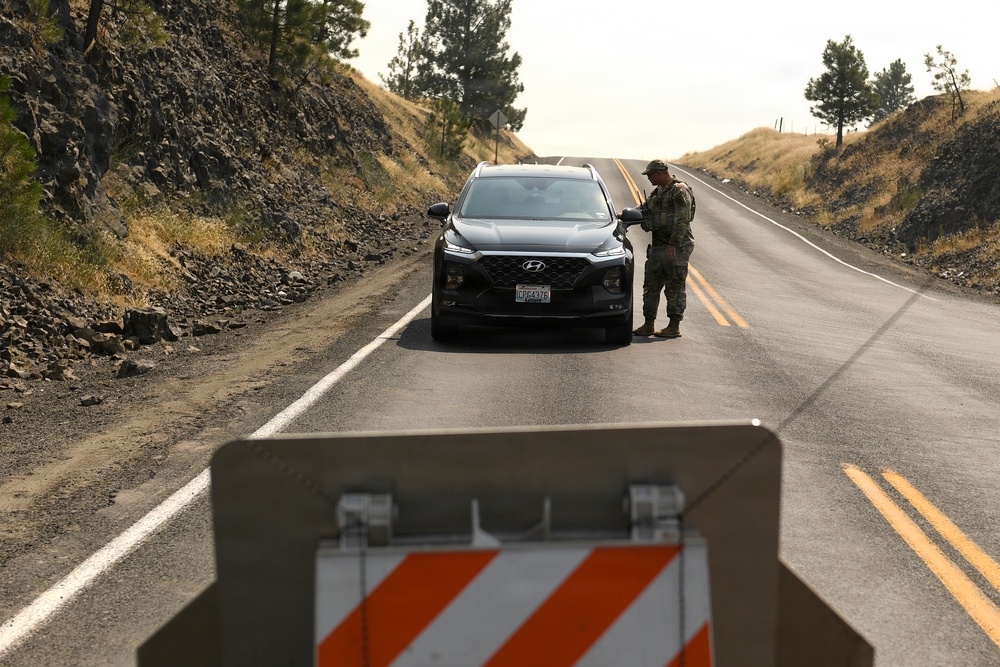 A military or law enforcement personnel in camouflage uniform is conducting a vehicle checkpoint stop on a two-lane rural road. A dark Hyundai SUV has been stopped at the checkpoint, with the officer standing at the driver's side window. A road barricade with orange and white diagonal stripes is visible in the foreground.