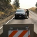 A military or law enforcement personnel in camouflage uniform is conducting a vehicle checkpoint stop on a two-lane rural road. A dark Hyundai SUV has been stopped at the checkpoint, with the officer standing at the driver's side window. A road barricade with orange and white diagonal stripes is visible in the foreground.