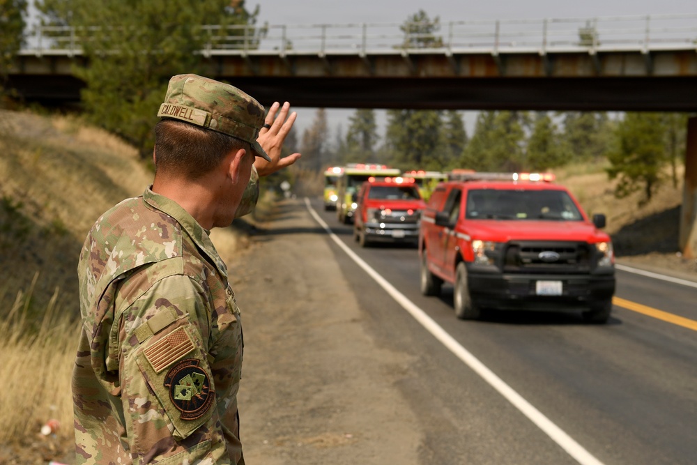 A U.S. military service member in camouflage uniform salutes a convoy of red fire department vehicles and emergency trucks passing on a two-lane highway. The scene appears to take place in a wildfire-affected area, evidenced by hazy smoke in the sky and scorched vegetation along the roadside. The gesture appears to be a show of respect toward the firefighting personnel.