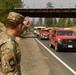 A U.S. military service member in camouflage uniform salutes a convoy of red fire department vehicles and emergency trucks passing on a two-lane highway. The scene appears to take place in a wildfire-affected area, evidenced by hazy smoke in the sky and scorched vegetation along the roadside. The gesture appears to be a show of respect toward the firefighting personnel.