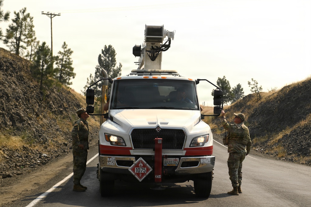 Two individuals in military camouflage uniforms are standing on either side of a large white utility/bucket truck stopped on a rural road. The truck displays a hazardous materials placard (UN 1075, flammable gas) and has a boom/crane apparatus mounted on top. The setting appears to be a hilly, semi-arid landscape with pine trees and rocky terrain.