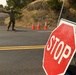 A military or law enforcement personnel in camouflage uniform is directing traffic at a road checkpoint. A portable stop sign is prominently placed in the foreground on a two-lane road, with orange traffic cones visible in the background. The setting appears to be a rural or semi-rural area with dry vegetation and trees on a hillside.