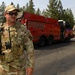 A U.S. Air Force Security Forces (SF) member in OCP (Operational Camouflage Pattern) uniform stands on a rural road holding a Motorola radio, wearing sunglasses and a tactical vest. Behind him is a large red fire truck with 'FIRE' lettered on its side, positioned on the road surrounded by pine trees. The scene suggests a joint military and fire department response operation, possibly related to wildfire management.