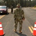 Military or law enforcement personnel in camouflage uniforms are manning a road checkpoint, stopping vehicles for inspection. Two personnel are visible, with one standing between traffic cones in the foreground and another approaching a dark red minivan. The surrounding area shows scorched or fire-damaged trees, suggesting this checkpoint may be related to wildfire emergency response or evacuation control.