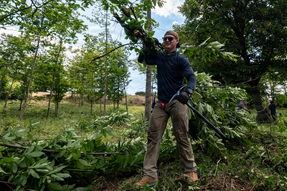A man is actively engaged in land clearing or vegetation management work, carrying cut branches and holding a long-handled tool. He is dressed in work attire including gloves, sunglasses, and a beanie, surrounded by piles of freshly cut leafy branches. The setting appears to be a wooded or semi-wooded outdoor area on an overcast but bright day.