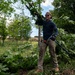 A man is actively engaged in land clearing or vegetation management work, carrying cut branches and holding a long-handled tool. He is dressed in work attire including gloves, sunglasses, and a beanie, surrounded by piles of freshly cut leafy branches. The setting appears to be a wooded or semi-wooded outdoor area on an overcast but bright day.