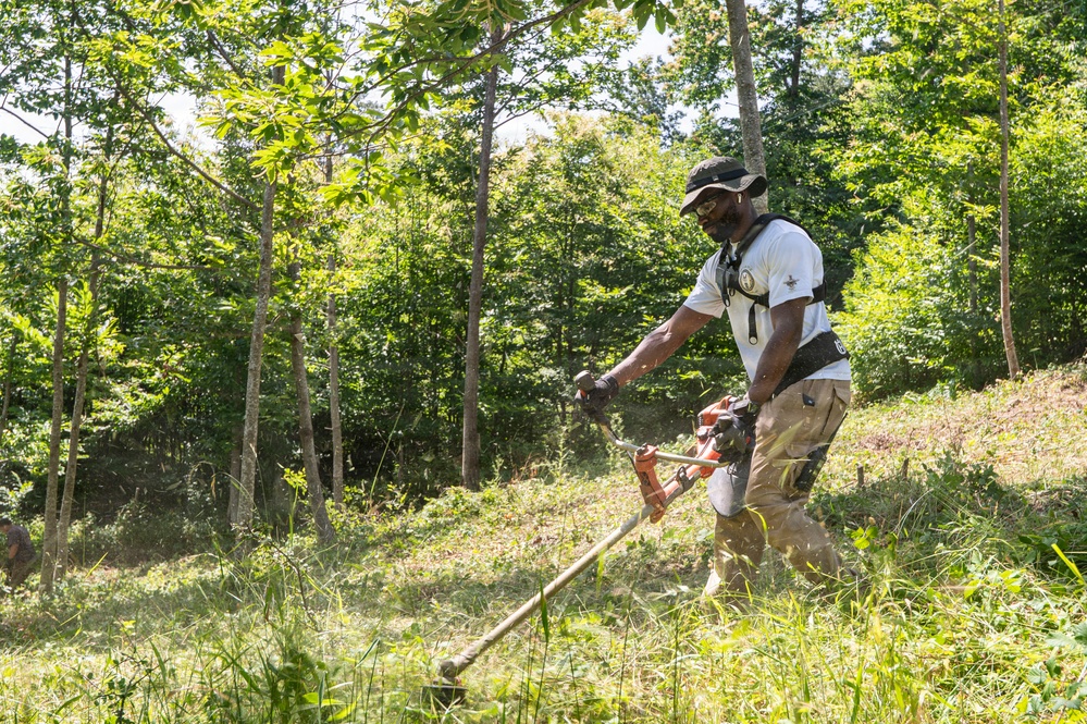 A man is operating a gas-powered string trimmer/brush cutter on a grassy hillside surrounded by dense green trees. He is leaning forward in an active working posture, clearing overgrown vegetation on what appears to be a sloped terrain. The image is taken outdoors on a bright, sunny day in a wooded or semi-wooded area.