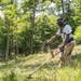 A man is operating a gas-powered string trimmer/brush cutter on a grassy hillside surrounded by dense green trees. He is leaning forward in an active working posture, clearing overgrown vegetation on what appears to be a sloped terrain. The image is taken outdoors on a bright, sunny day in a wooded or semi-wooded area.