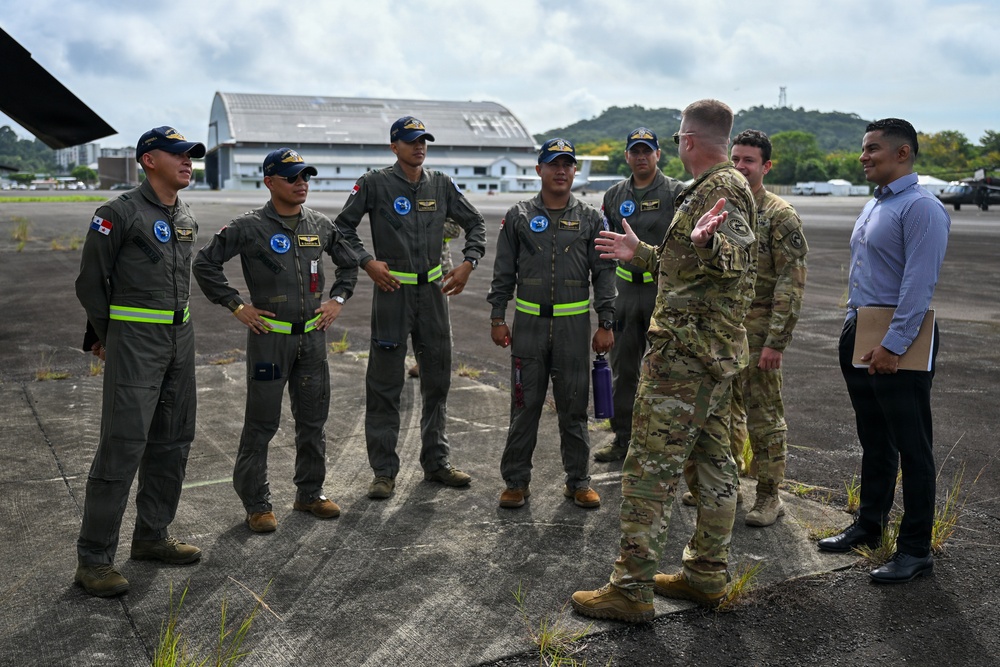 A U.S. Army soldier in MultiCam/OCP camouflage uniform is briefing or conversing with a group of approximately five aviators wearing olive-green flight suits with reflective yellow-green striping and blue caps bearing wing insignia. At least one flight suit displays a Panamanian flag patch, suggesting this is a bilateral military engagement at an airfield. A civilian interpreter or liaison holding a folder stands to the right of the U.S. soldier.