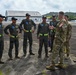 A U.S. Army soldier in MultiCam/OCP camouflage uniform is briefing or conversing with a group of approximately five aviators wearing olive-green flight suits with reflective yellow-green striping and blue caps bearing wing insignia. At least one flight suit displays a Panamanian flag patch, suggesting this is a bilateral military engagement at an airfield. A civilian interpreter or liaison holding a folder stands to the right of the U.S. soldier.