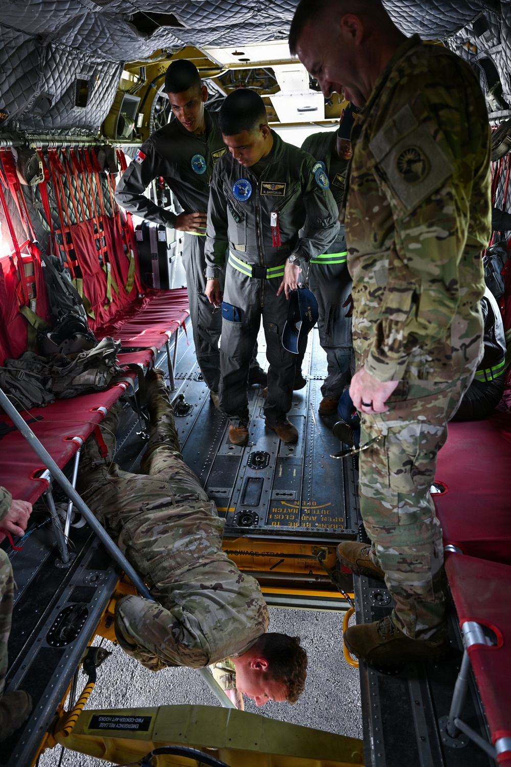 Military personnel are gathered inside what appears to be a large military transport aircraft, likely a C-130 or similar cargo/transport aircraft. Personnel in both camouflage (OCP/MultiCam) uniforms and dark olive flight suits are examining or demonstrating litter/stretcher equipment on the aircraft floor. This appears to be a training or familiarization exercise, possibly involving aeromedical evacuation procedures.