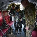 Military personnel are gathered inside what appears to be a large military transport aircraft, likely a C-130 or similar cargo/transport aircraft. Personnel in both camouflage (OCP/MultiCam) uniforms and dark olive flight suits are examining or demonstrating litter/stretcher equipment on the aircraft floor. This appears to be a training or familiarization exercise, possibly involving aeromedical evacuation procedures.