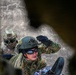 Two military personnel are visible in a tactical environment, photographed through a gap or opening in a structure. The foreground soldier wears a green FAST-style helmet, sunglasses, black tactical gloves, and a digital camouflage uniform, with one hand raised. A second soldier in the background wears a multicam uniform with a helmet and protective goggles.