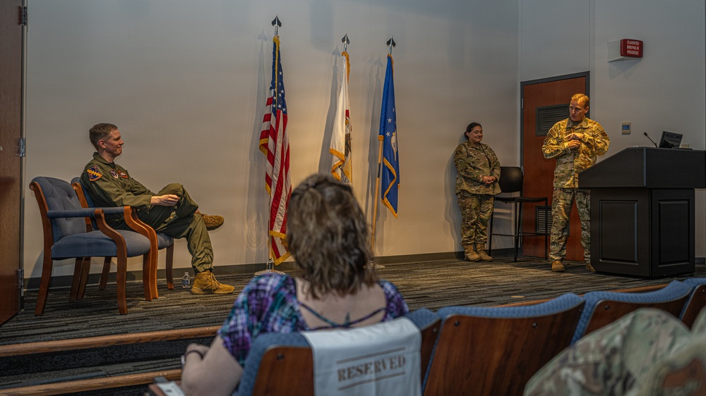 A military briefing or ceremony is taking place in what appears to be an auditorium or conference room on a military installation. Three military personnel are on a stage area, with one seated in a flight suit and two others in Army Combat Uniforms (ACU/OCP), one of whom is speaking near a podium. A civilian audience member is visible in the foreground seated in a reserved seat.