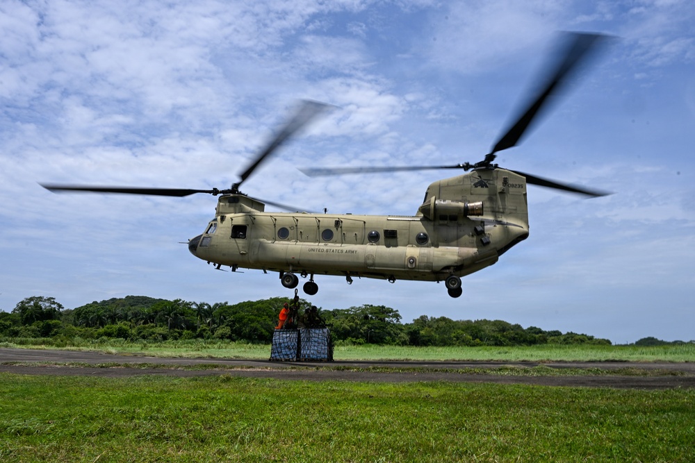 A United States Army CH-47 Chinook helicopter hovers low over a grassy field while personnel on the ground attach or secure a pallet of supplies beneath it. The scene appears to depict a sling-load or humanitarian aid delivery operation in a tropical or subtropical environment. The helicopter's dual rotors are in motion, creating visible blur.