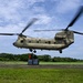 A United States Army CH-47 Chinook helicopter hovers low over a grassy field while personnel on the ground attach or secure a pallet of supplies beneath it. The scene appears to depict a sling-load or humanitarian aid delivery operation in a tropical or subtropical environment. The helicopter's dual rotors are in motion, creating visible blur.