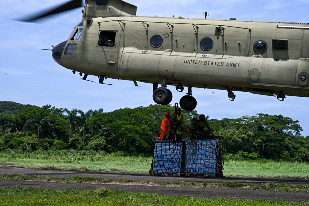 A United States Army CH-47 Chinook helicopter hovers low over a tarmac area surrounded by tropical vegetation. Ground personnel, including one individual in a high-visibility orange vest and others in military camouflage, are working to attach or secure a netted pallet of supplies — appearing to be bottled water — to the helicopter for a sling-load operation. This appears to be a humanitarian aid or disaster relief delivery mission.