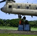 A United States Army CH-47 Chinook helicopter hovers low over a tarmac area surrounded by tropical vegetation. Ground personnel, including one individual in a high-visibility orange vest and others in military camouflage, are working to attach or secure a netted pallet of supplies — appearing to be bottled water — to the helicopter for a sling-load operation. This appears to be a humanitarian aid or disaster relief delivery mission.
