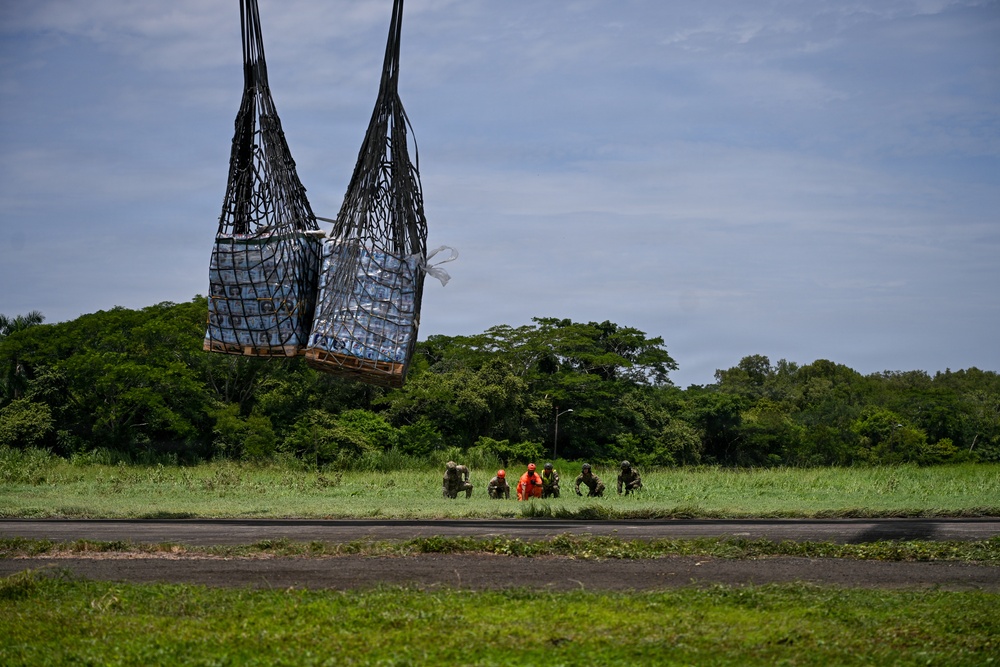 A helicopter sling-load operation is underway at what appears to be a remote airstrip or landing zone surrounded by dense tropical vegetation. A large cargo net containing palletized supplies, likely bottled water or humanitarian aid, is suspended in the air. Several personnel, including at least one in an orange vest, are crouched on the ground nearby, likely serving as ground guides for the landing.