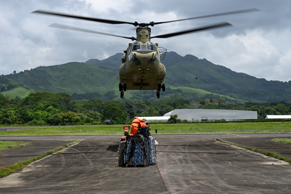 A military CH-47 Chinook helicopter hovers low over a tarmac while ground personnel in orange vests prepare a netted cargo load for sling-load extraction. The operation appears to be a humanitarian aid or logistics mission, with blue-wrapped pallets secured in a cargo net on the ground. The setting is a tropical airfield surrounded by lush green mountains under overcast skies.