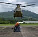 A military CH-47 Chinook helicopter hovers low over a tarmac while ground personnel in orange vests prepare a netted cargo load for sling-load extraction. The operation appears to be a humanitarian aid or logistics mission, with blue-wrapped pallets secured in a cargo net on the ground. The setting is a tropical airfield surrounded by lush green mountains under overcast skies.