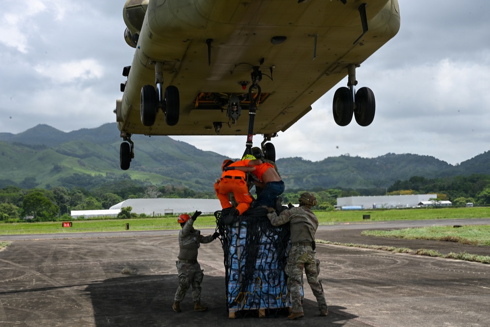 Military and civilian personnel are attaching a cargo net-wrapped pallet of supplies to the underside of a large military helicopter, likely a CH-47 Chinook, on a tarmac. The operation appears to be a sling-load or external lift preparation for humanitarian aid delivery. The lush green mountainous terrain in the background suggests a tropical location.
